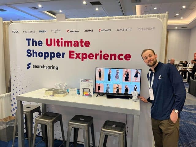 Casey Marksberry, Searchspring Sales Rep at eTailWest 2023 Searchspring booth. "The Ultimate Shopper Experience" is in bold letters on the backdrop followed by the Searchspring logo. Brand logos line the top of the backdrop. A table with 4 chairs is against the backdrop with a computer showing a video and table signs.