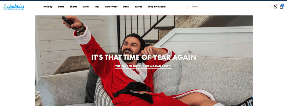 Chubbies holiday collection promotion banner - white, 20-something male with beard in a red and white santa robe, sitting on couch pointing